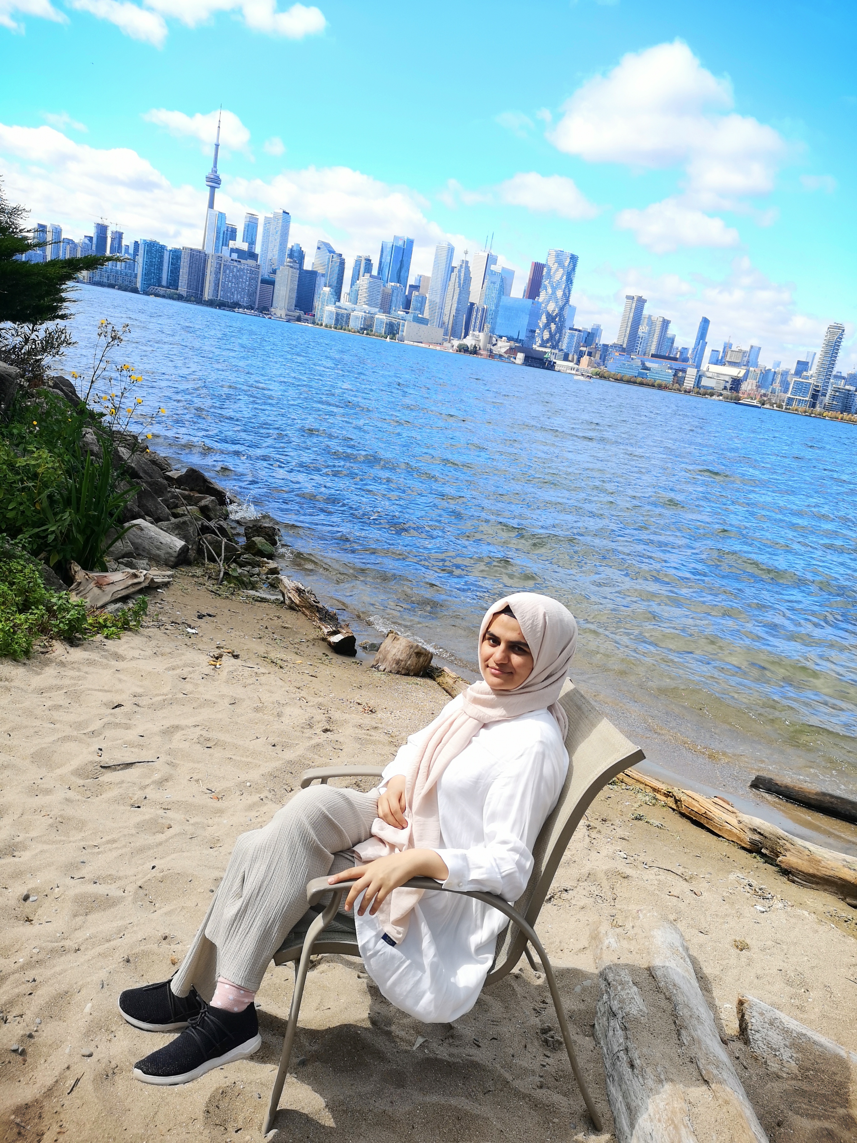 Berin sitting on a chair on the beach with buildings of Toronto behind her