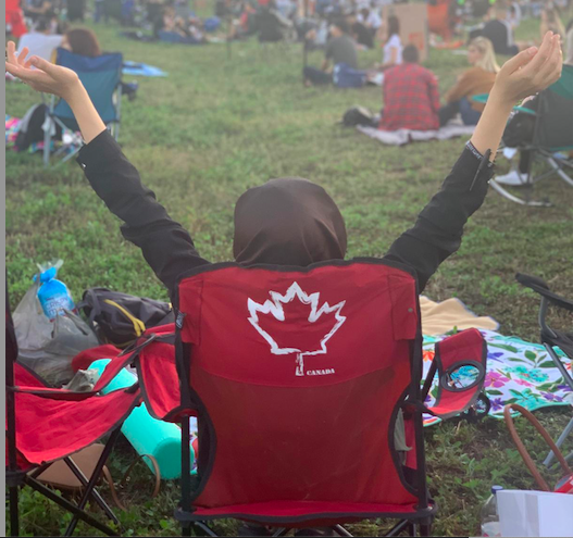 Berin sitting in a camping chair back-on with a maple leaf on the back of it in a field of people watching a concert